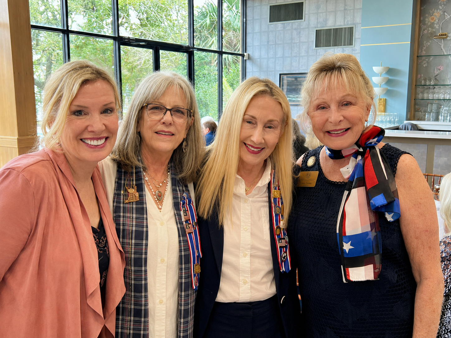 Four women standing close together, smiling, indoors at a social event. They are dressed in business casual attire with patriotic-themed ribbons and pins, and one woman has a scarf with American flag colors. Background shows large windows, trees outside, and a modern interior setting.