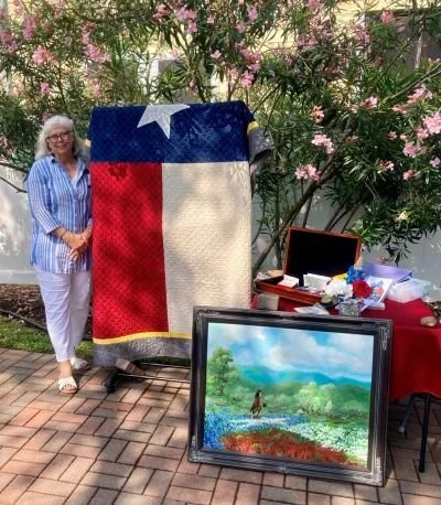 DRT member standing outdoors next to a Texas flag quilt display and a table with Texas items, with flowering bushes in the background.
