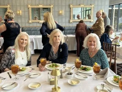 Three women sitting at a table at the Artillery Club in Galveston, enjoying the DRT Texas History Lunch, with other people in the background.