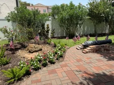 The Cradle backyard garden with green bushes, flowering plants, and small American flags, brick patio, and a 1812 cannon on the patio.