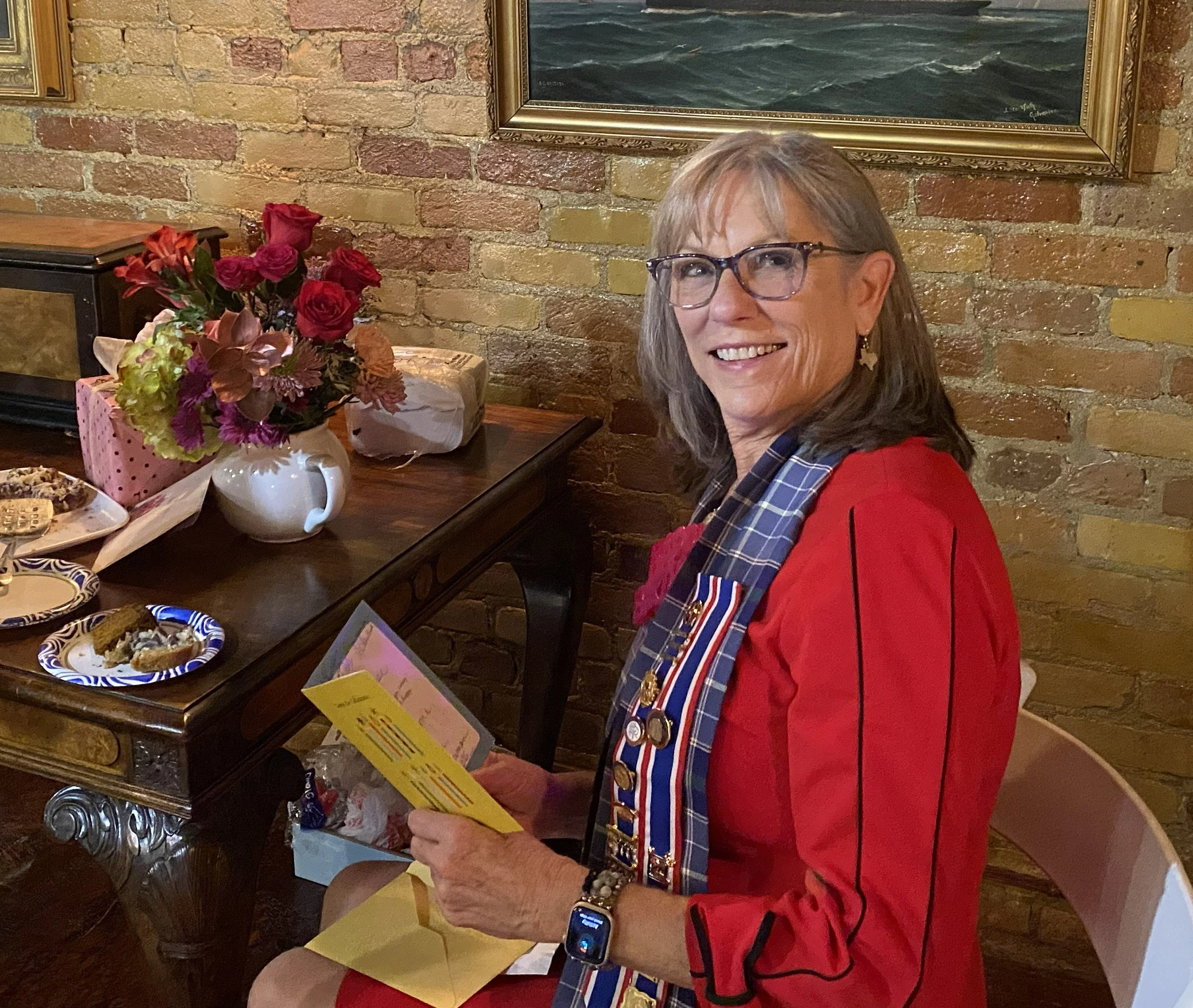 DRT member with gray hair and glasses smiling, wearing a red jacket and plaid sash with medals, sitting next to a table with a bouquet of flowers, a card, and snacks, at the Wine Room of the Bryan Museum in Galveston.
