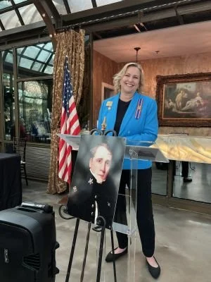 DRT member in a blue blazer speaking at a podium in the Conservatory of the Bryan Museum in Galveston, with a portrait of Sidney Sherman on an easel beside her and an American flag in the background.