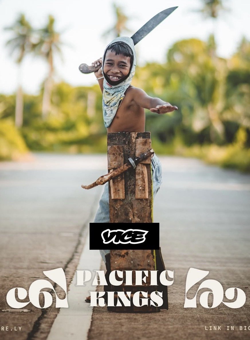 Smiling young boy with a bandana around his head, shirtless, standing behind a cardboard and wood shield, with a machete in one hand, on a paved road with greenery and palm trees in the background.