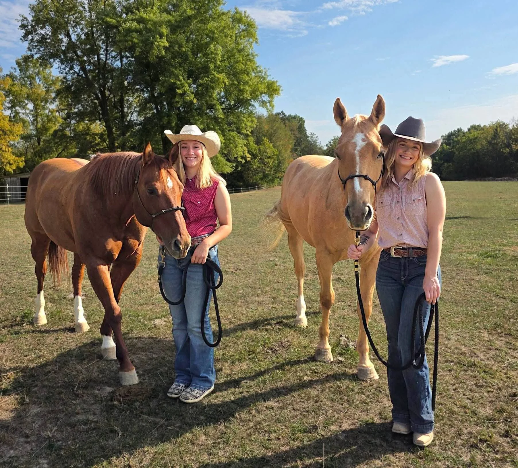 Two young women smiling and standing with horses on a grassy field. One woman is wearing a white cowboy hat, a pink sleeveless top, and blue jeans. The other woman is wearing a gray cowboy hat, a light pink sleeveless top, and dark jeans. The horses 