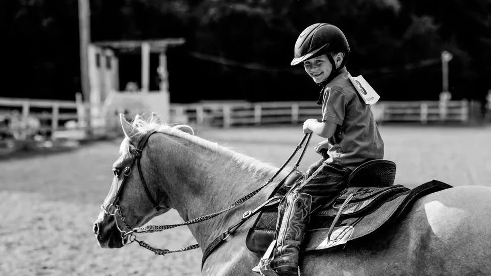 A young boy wearing a helmet and a shirt, smiling as he rides a riding horse in an outdoor arena.