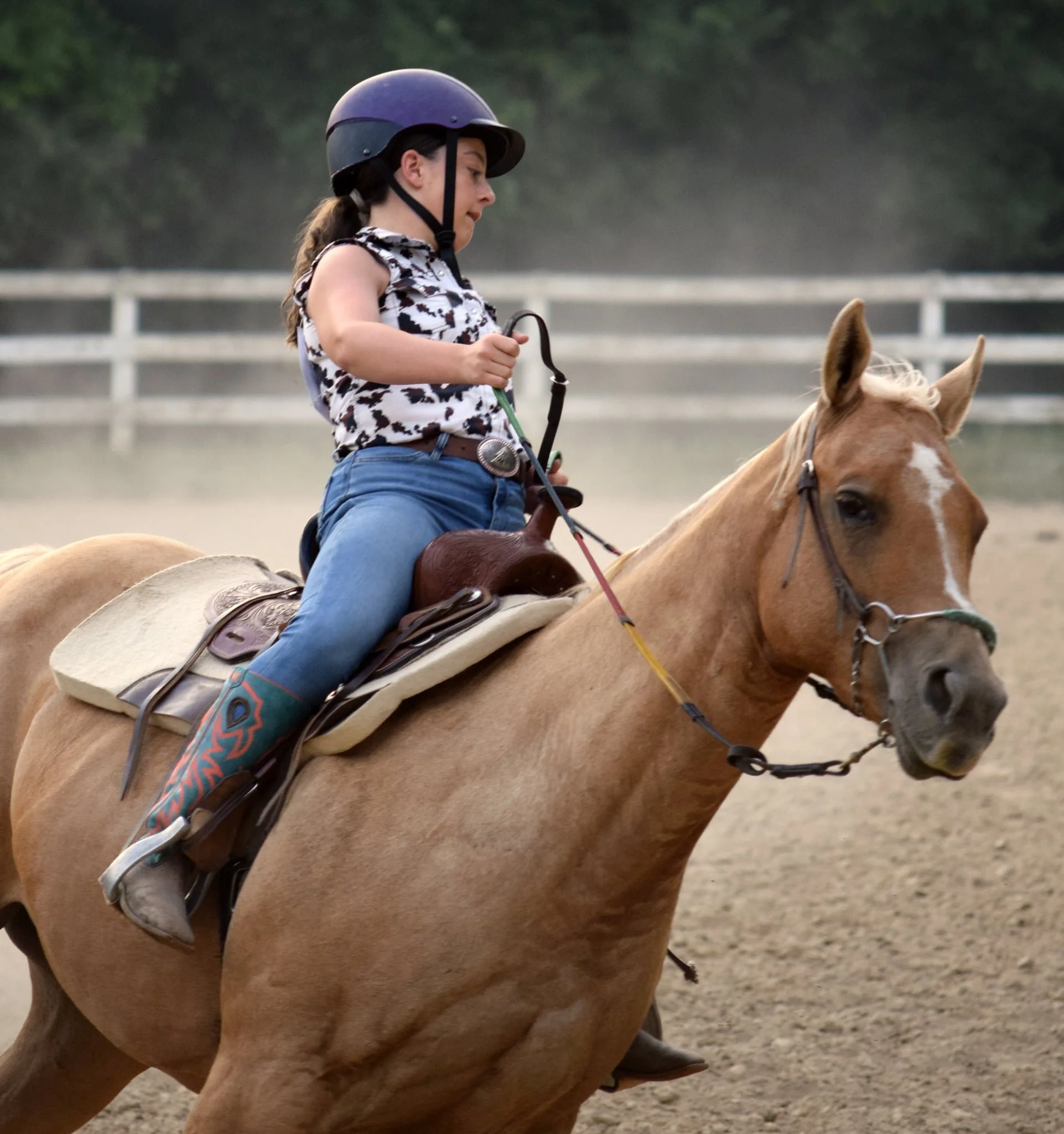 A young girl riding a horse during daytime, wearing a helmet and Western-style clothing, with a white fence and out-of-focus trees in the background.