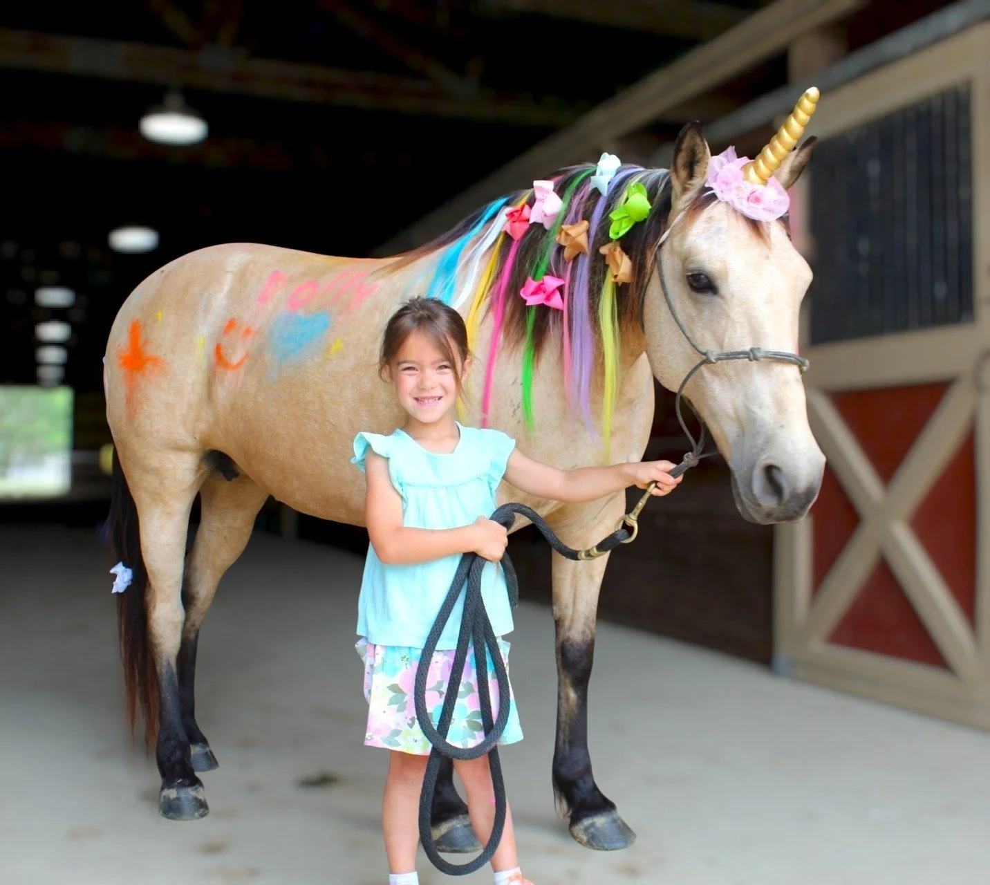 A young girl in a light blue dress smiling and holding the reins of a decorated unicorn horse inside a stable or riding arena.