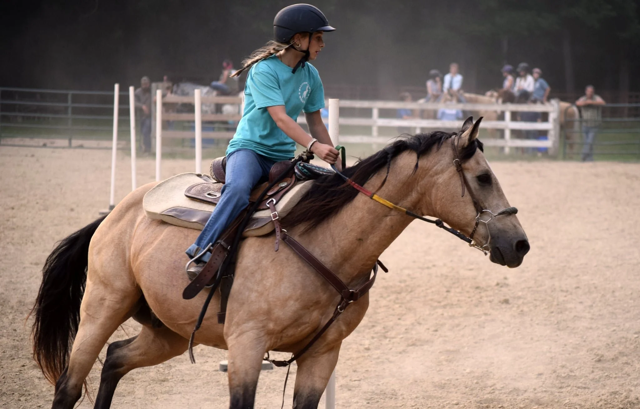 A young woman in a black riding helmet riding a light brown horse with dark mane in an outdoor arena during daytime, with several spectators in the background.