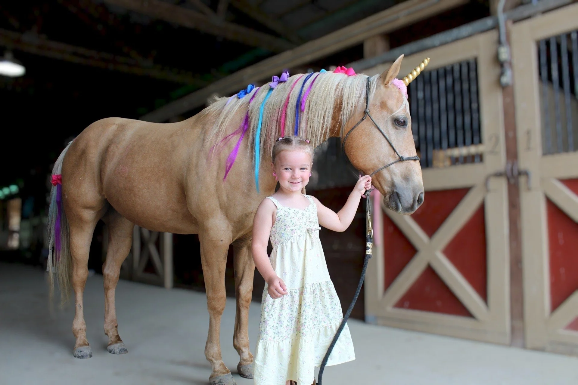 A young girl in a white dress standing next to a light brown horse with a rainbow-colored mane and horn, inside a stable.
