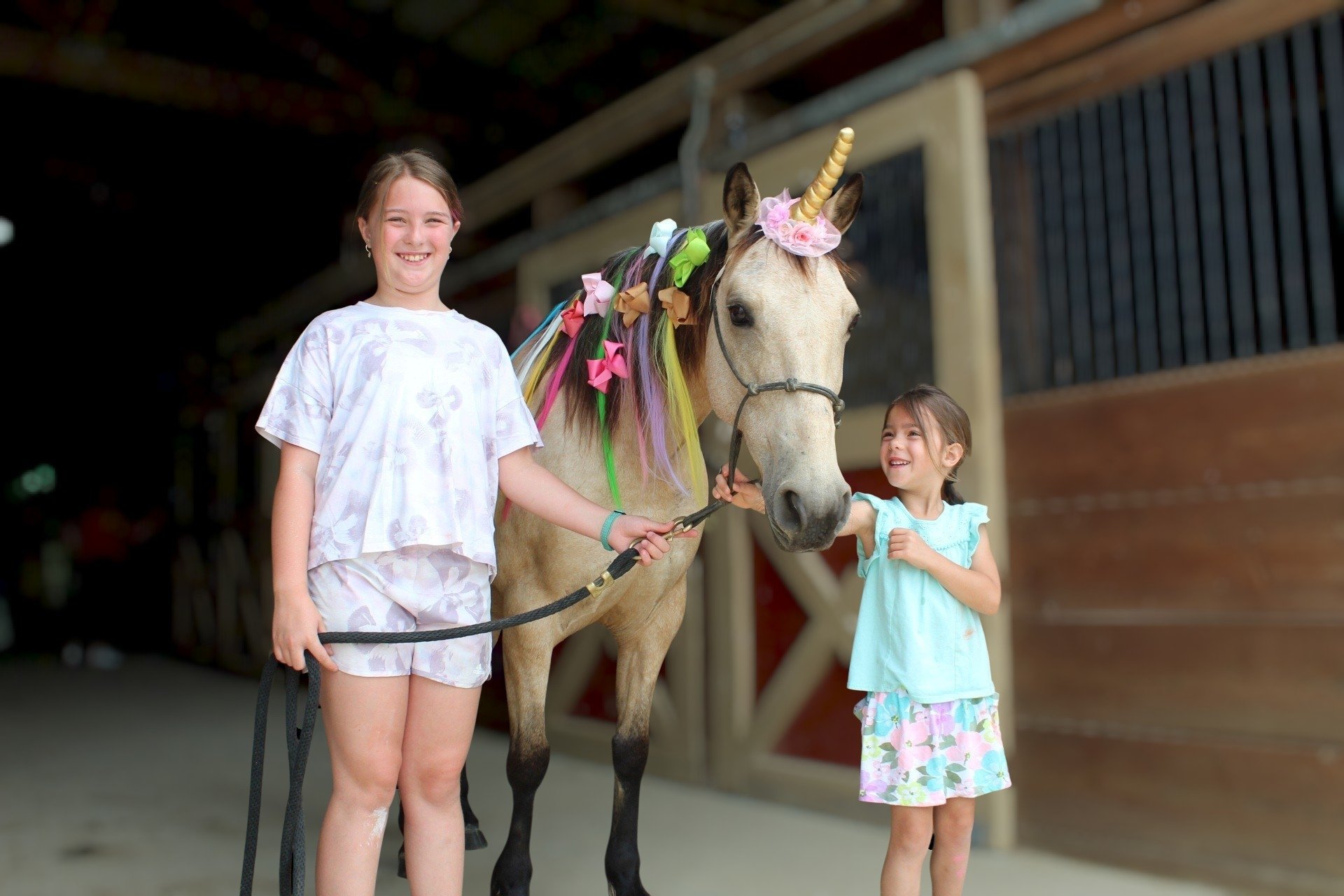 Two young girls and a unicorn horse standing in a barn. The horse is decorated with colorful ribbons and a horn with flowers, and both girls are smiling.
