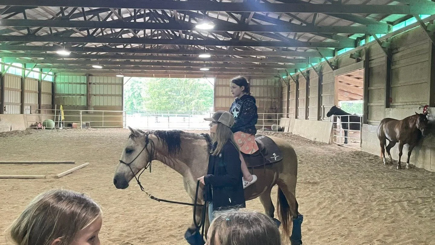 Children riding a horse in an indoor riding arena, guided by an instructor.