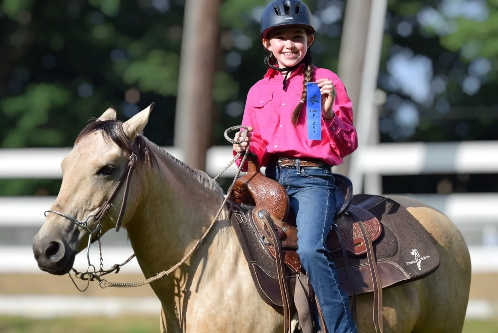 A young girl riding a horse at an equestrian event, smiling while holding a first place ribbon.
