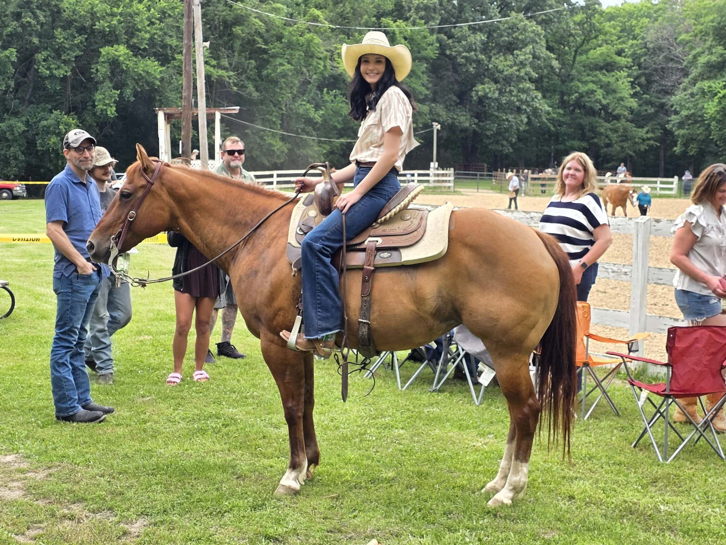 A young woman with long dark hair smiling while sitting on a brown horse, wearing a large sun hat and casual clothing, at an outdoor event with other people and horses in the background.