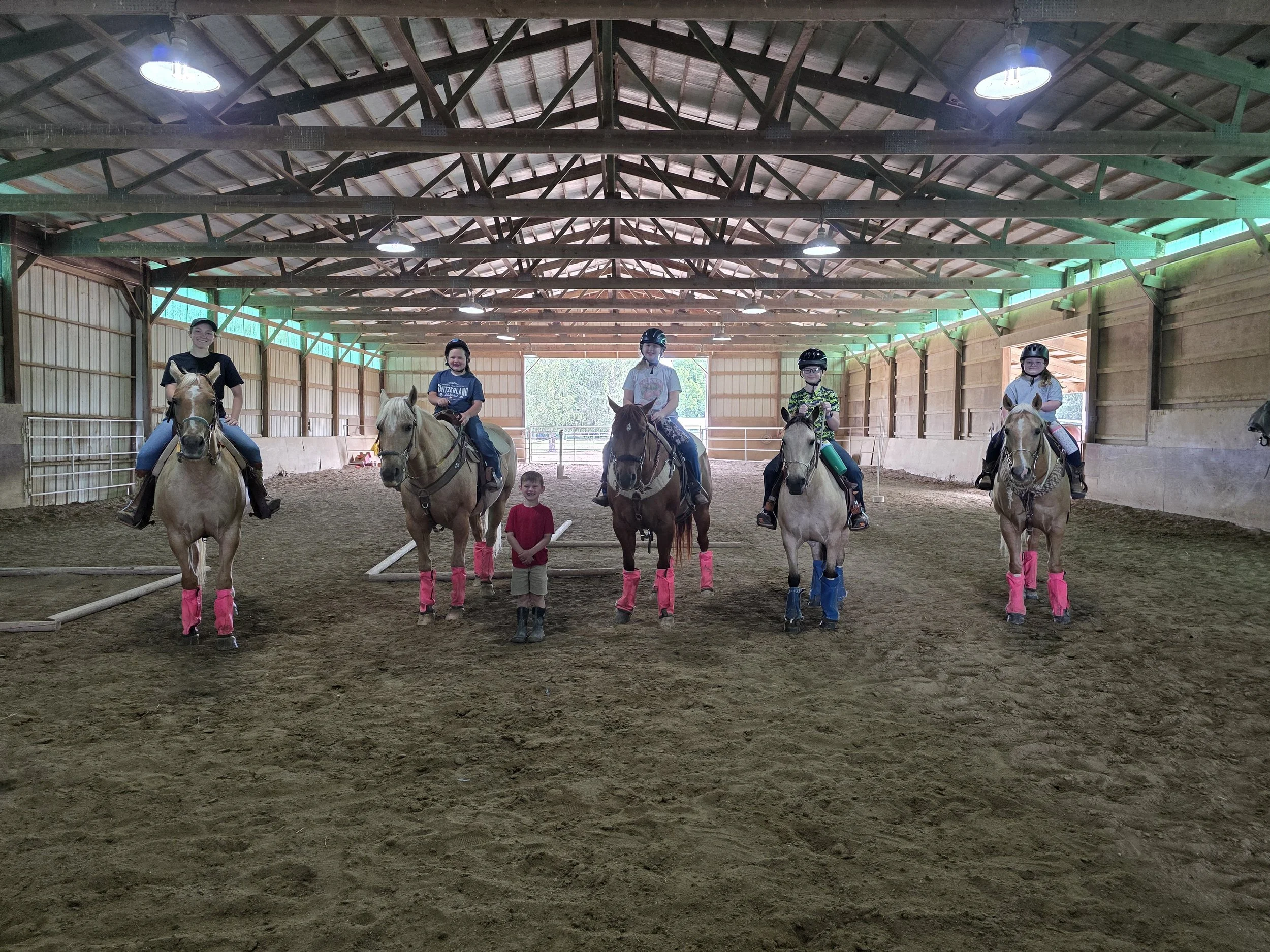 Five children riding horses inside a covered indoor riding arena. All children are wearing helmets, and most are wearing glasses. The arena has a high wooden ceiling with visible beams and lights. The children are smiling and appear to be enjoying th