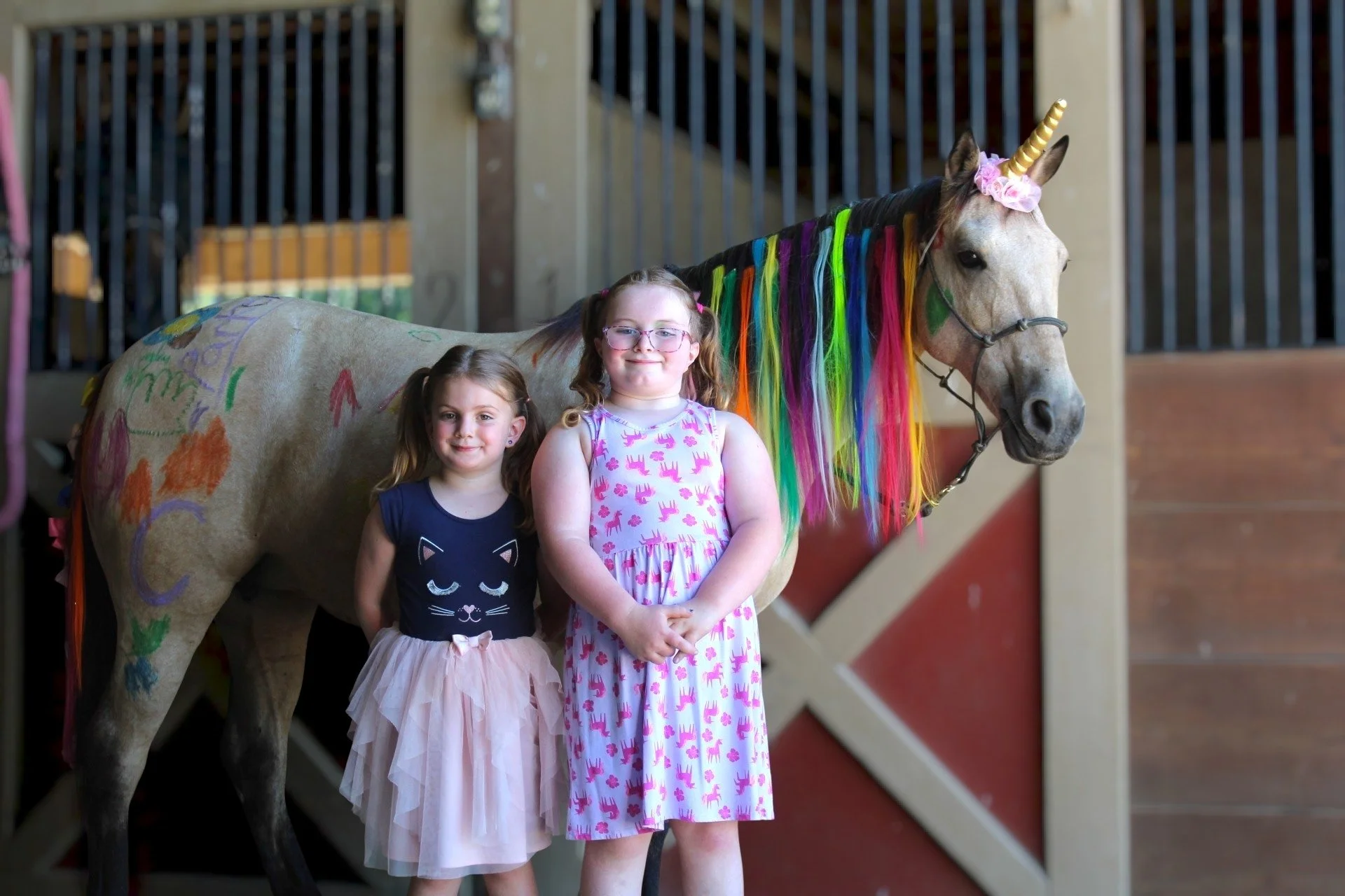Two young girls standing next to a painted unicorn at an indoor farm or petting zoo. The unicorn has a rainbow-colored mane and a gold horn, with colorful drawings on its body.