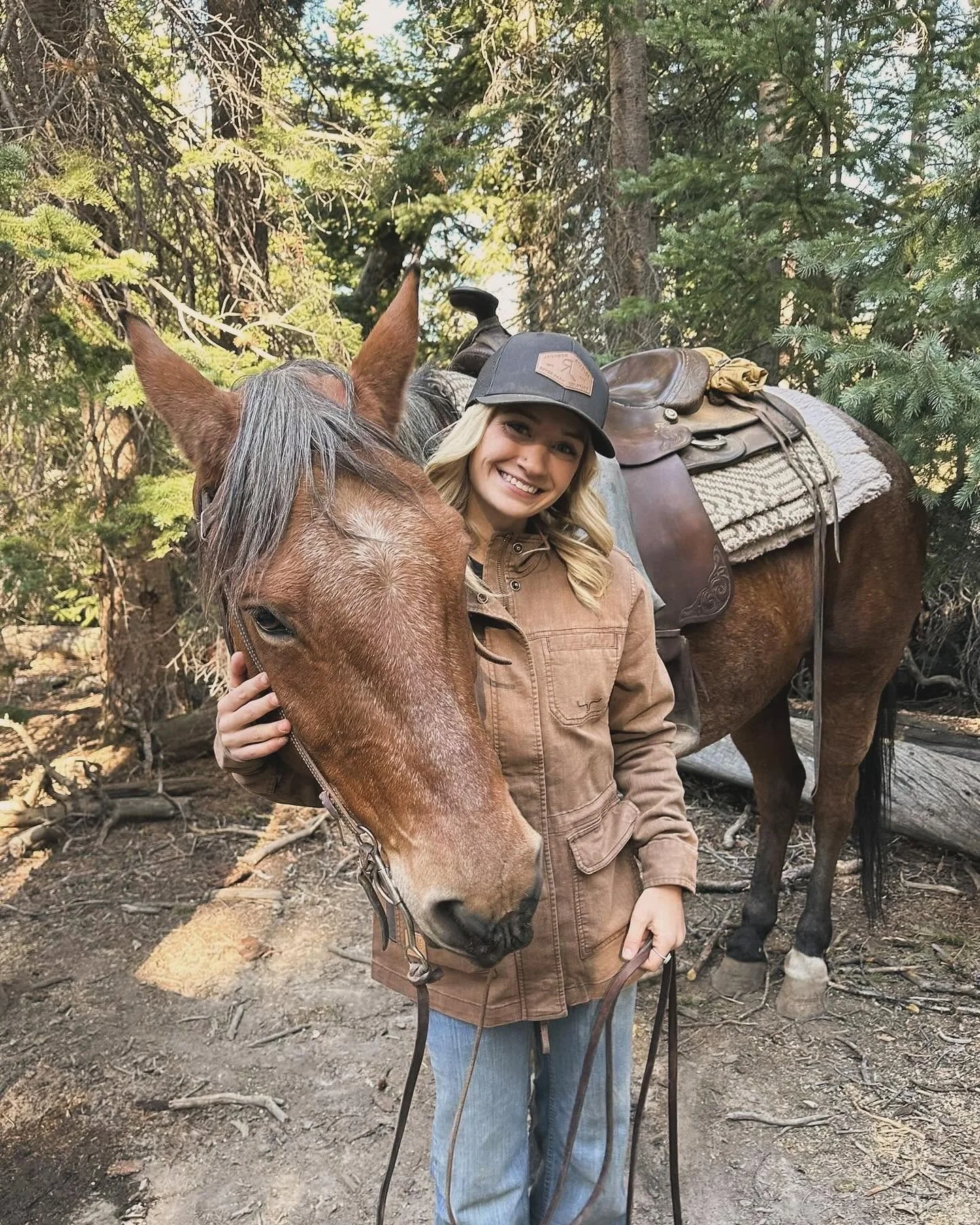 A woman smiling while holding a brown horse's bridle in a wooded outdoor setting. The horse has a saddle and blanket on its back, with trees surrounding them.