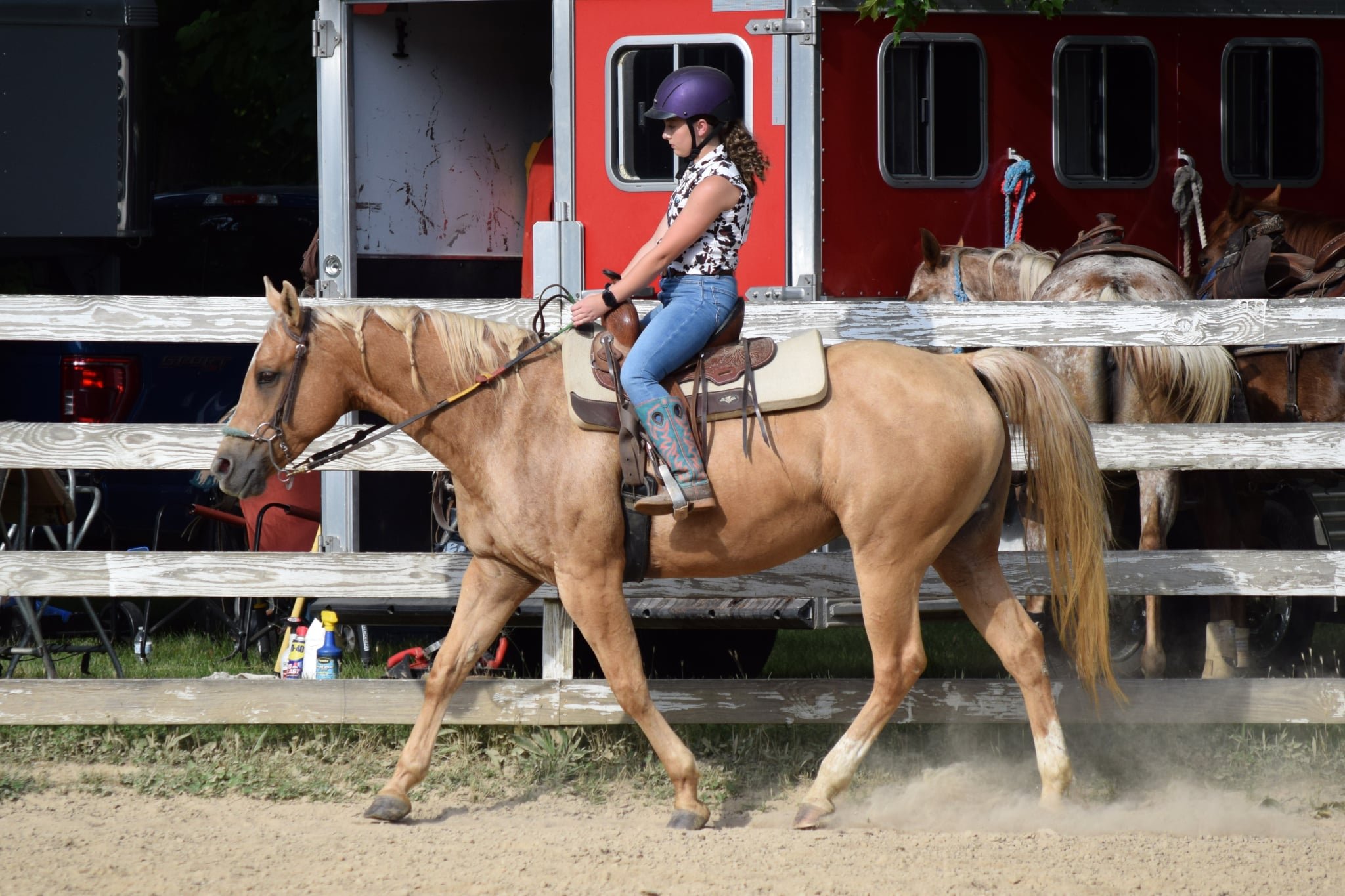 A young woman with curly hair wearing a purple riding helmet, a sleeveless floral shirt, and blue jeans, riding a tan horse with a light mane, on a dirt track. Behind her is a red horse trailer with several horses inside, some partially visible.