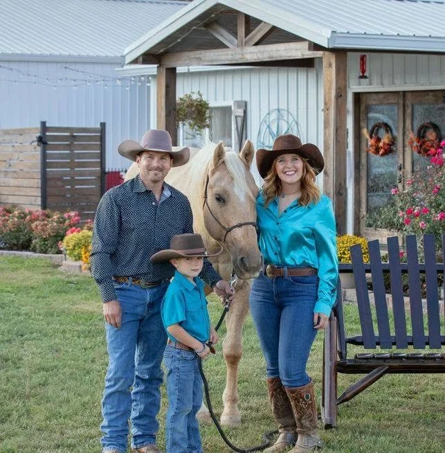 A family of three, including a man, woman, and child, with a light-colored horse, standing outside a farm building with flowers and decorations.