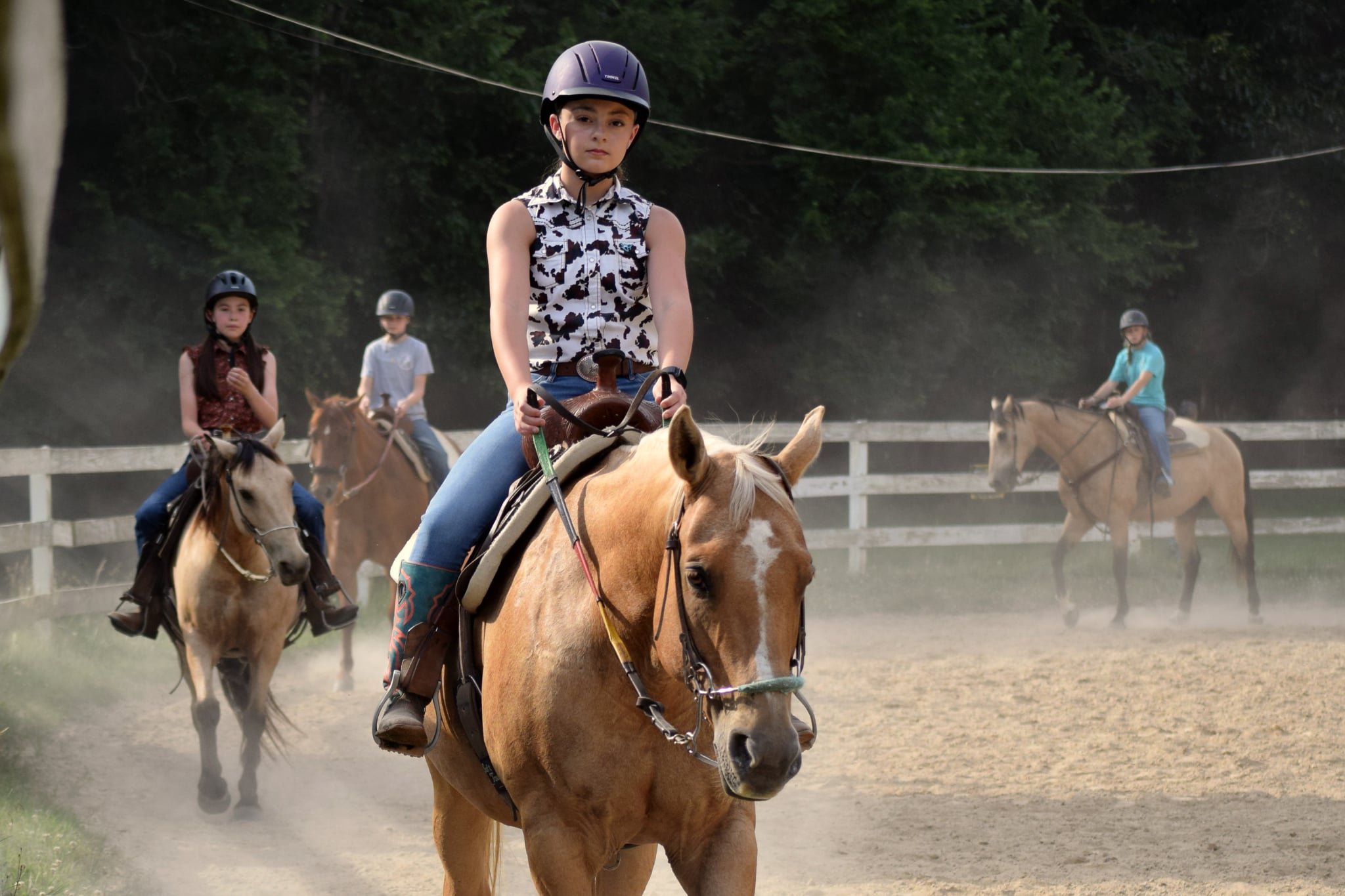 A young girl riding a horse in an outdoor riding arena with other children riding nearby, all wearing helmets.