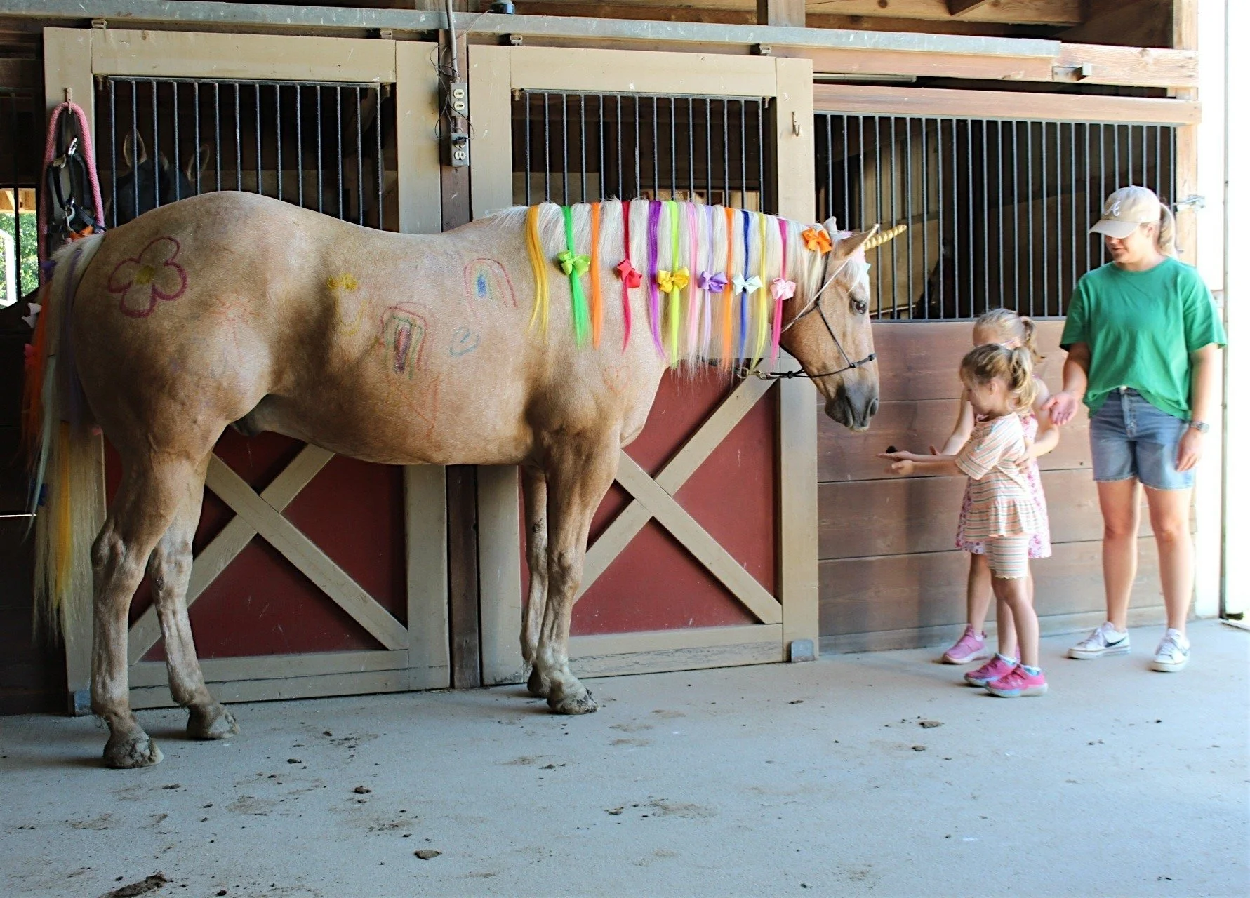 A tan horse with a rainbow-colored mane and tail, decorated with flower-shaped ribbons, standing in a barn with children and a woman nearby.