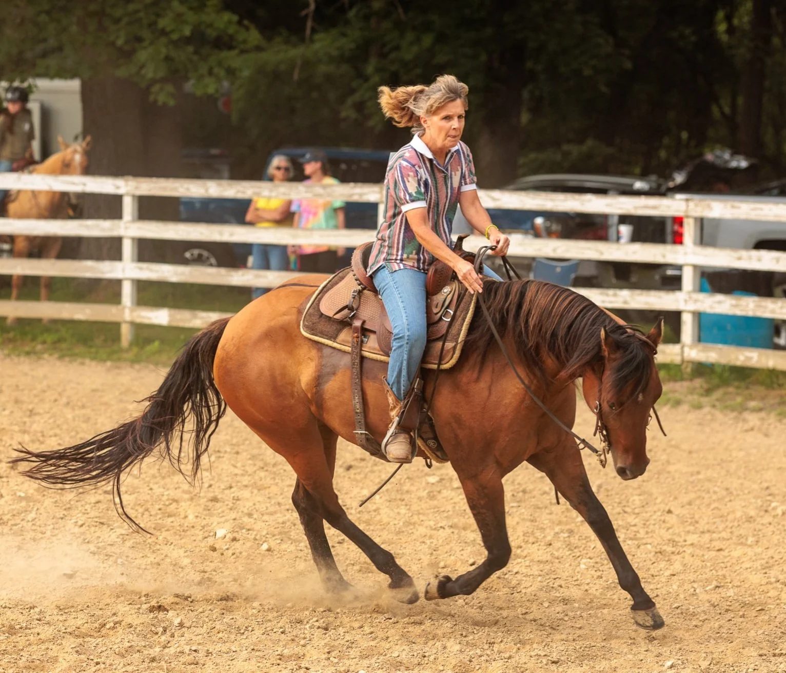 A woman riding a brown horse on a dirt track, wearing jeans and a multicolored striped shirt, with trees and parked cars in the background.