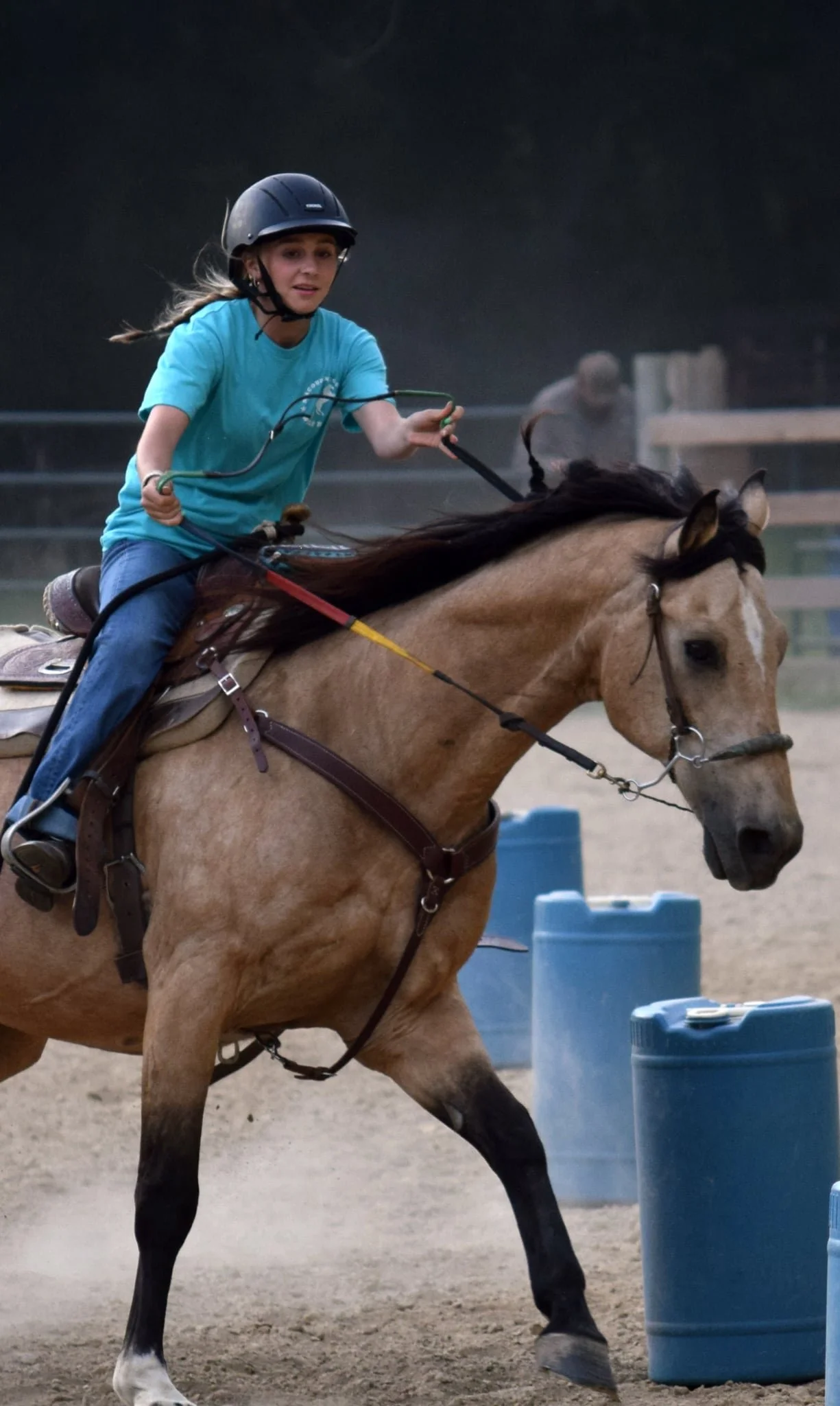 A girl wearing a helmet riding a galloping tan horse in an equestrian arena, with blue barrels in the background.