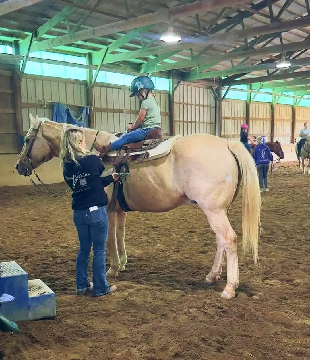 A girl is sitting on a light-colored horse inside an indoor riding arena, wearing a helmet and a green t-shirt. A woman is standing next to the horse, holding the saddle. Other people and horses can be seen in the background.