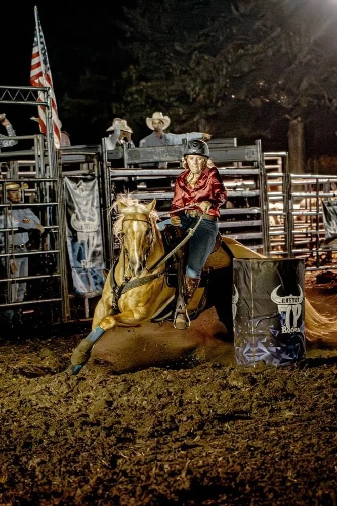 A woman riding a galloping horse during a rodeo at night, wearing a helmet and a red jacket, with cowboys and American flag in the background.