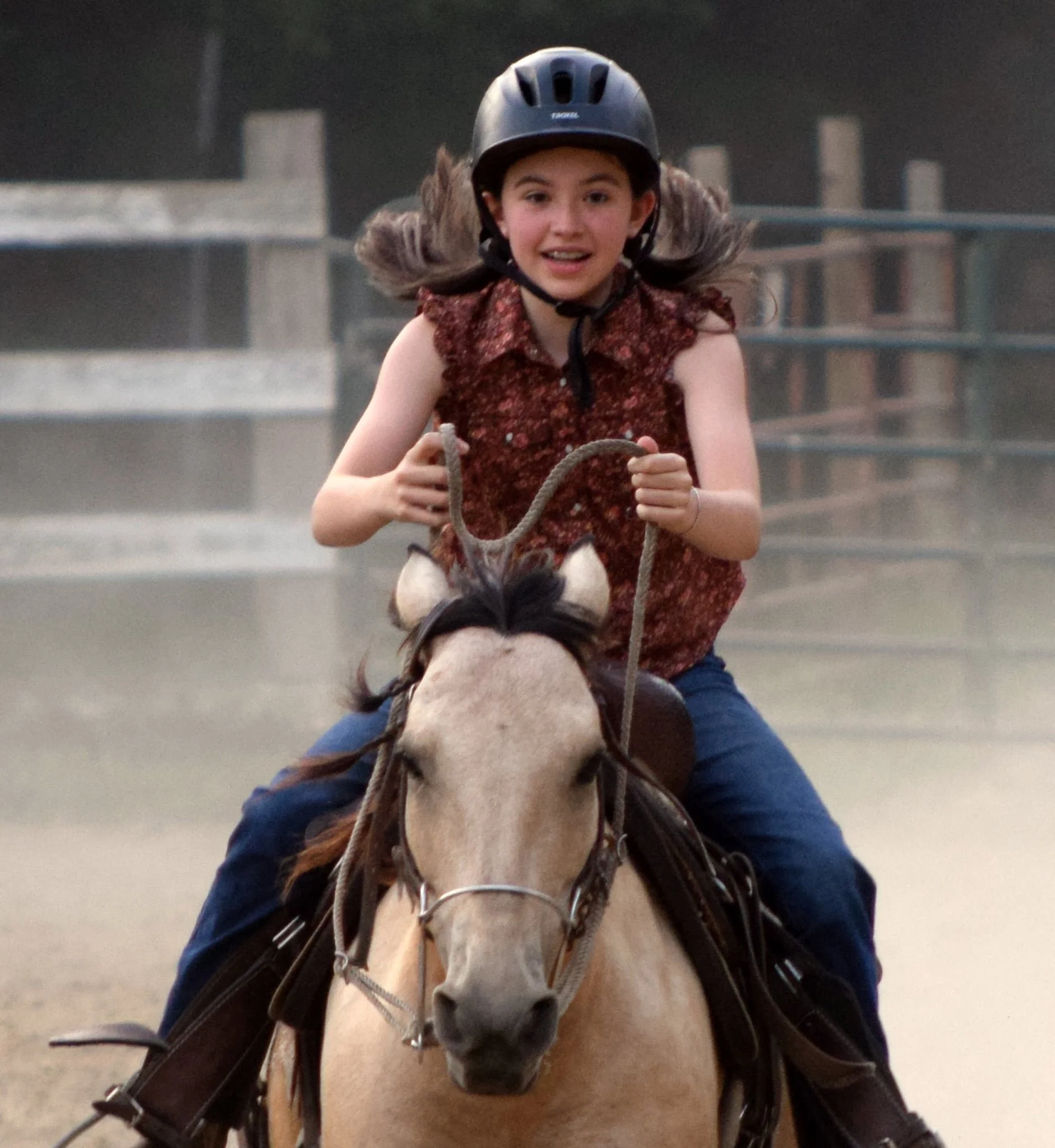 A young girl with long hair riding a light-colored horse, wearing a black riding helmet and a sleeveless patterned shirt, in an outdoor stable area.