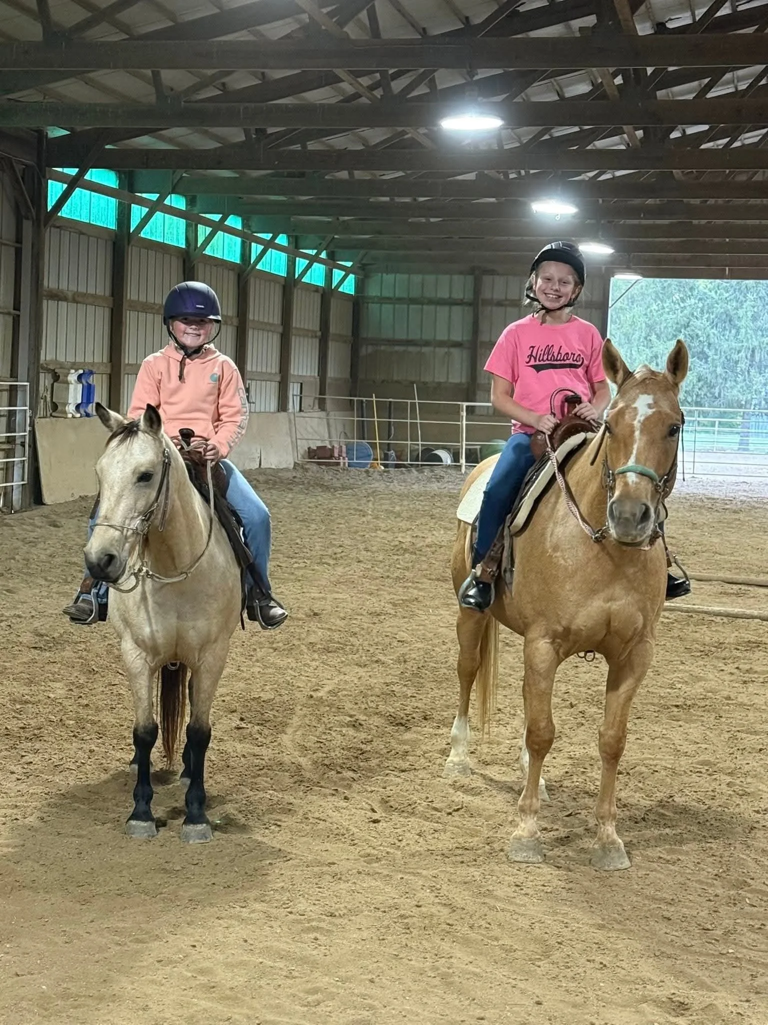 Two young children riding horses indoors in a riding arena, both wearing helmets, smiling.