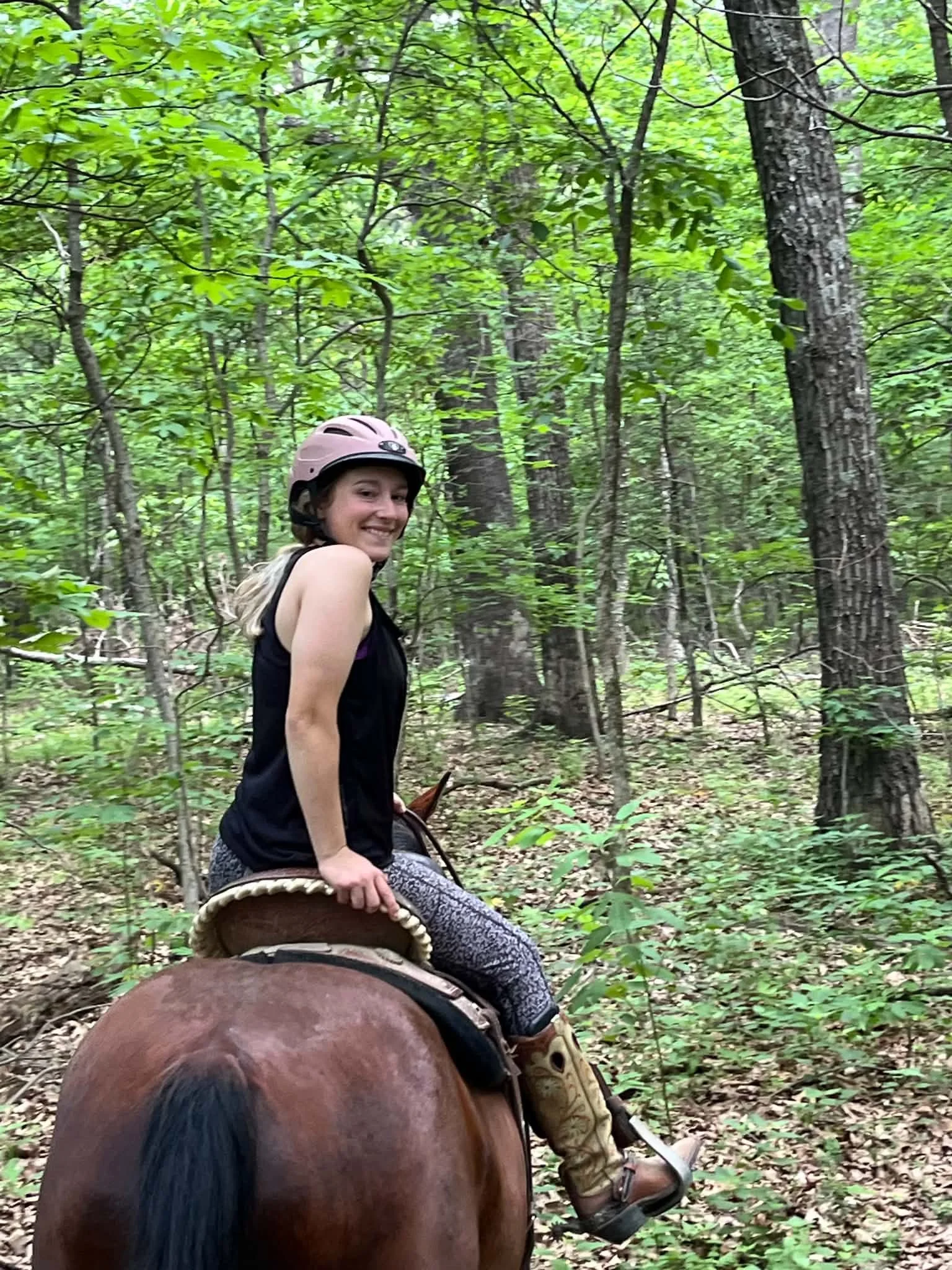 A young woman wearing a pink helmet and black sleeveless top riding a horse through a green forest, with leafy trees and ground covered with leaves.