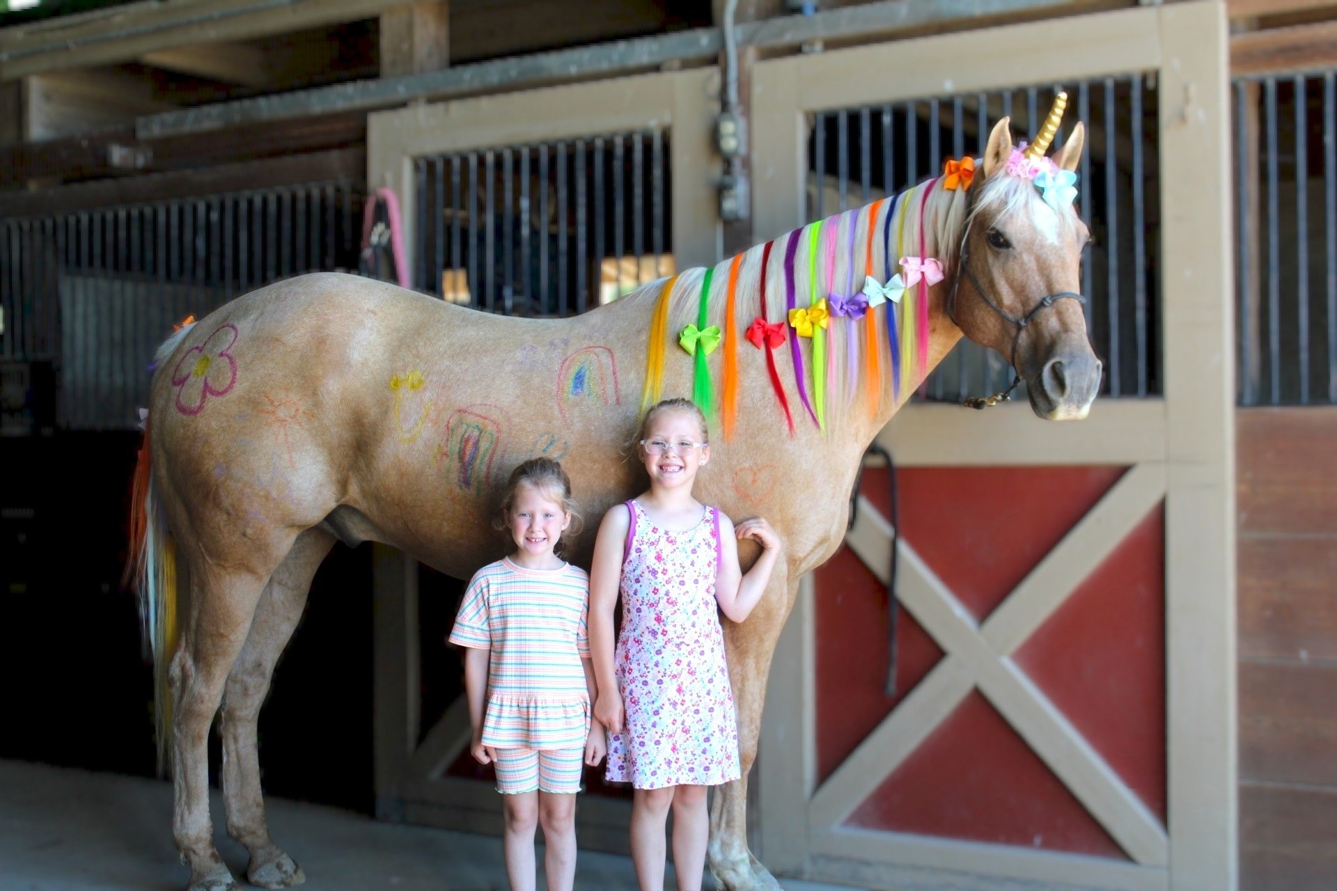 Two young girls are standing in front of a decorated unicorn statue inside a barn or stable. The unicorn has a rainbow-colored mane and tail, and is adorned with colorful bows, rainbow drawings, and a unicorn horn headband. The girls are smiling and 