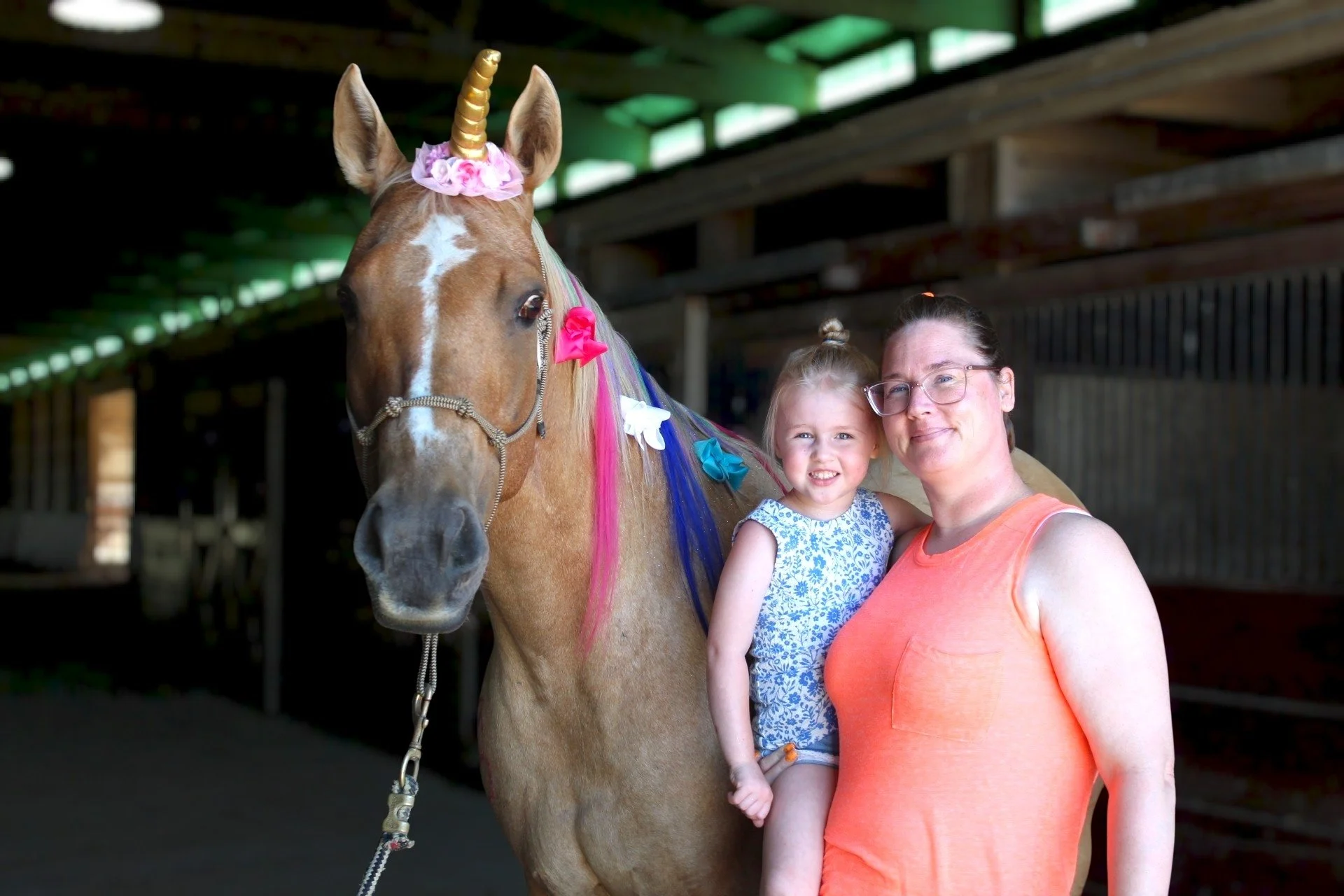 A woman and a young girl taking a selfie with a decorated unicorn horse in a barn.