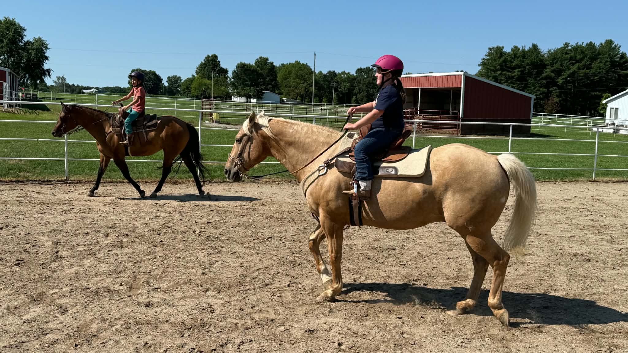 Two children riding horses in a fenced outdoor riding arena on a sunny day, with green fields and trees in the background.