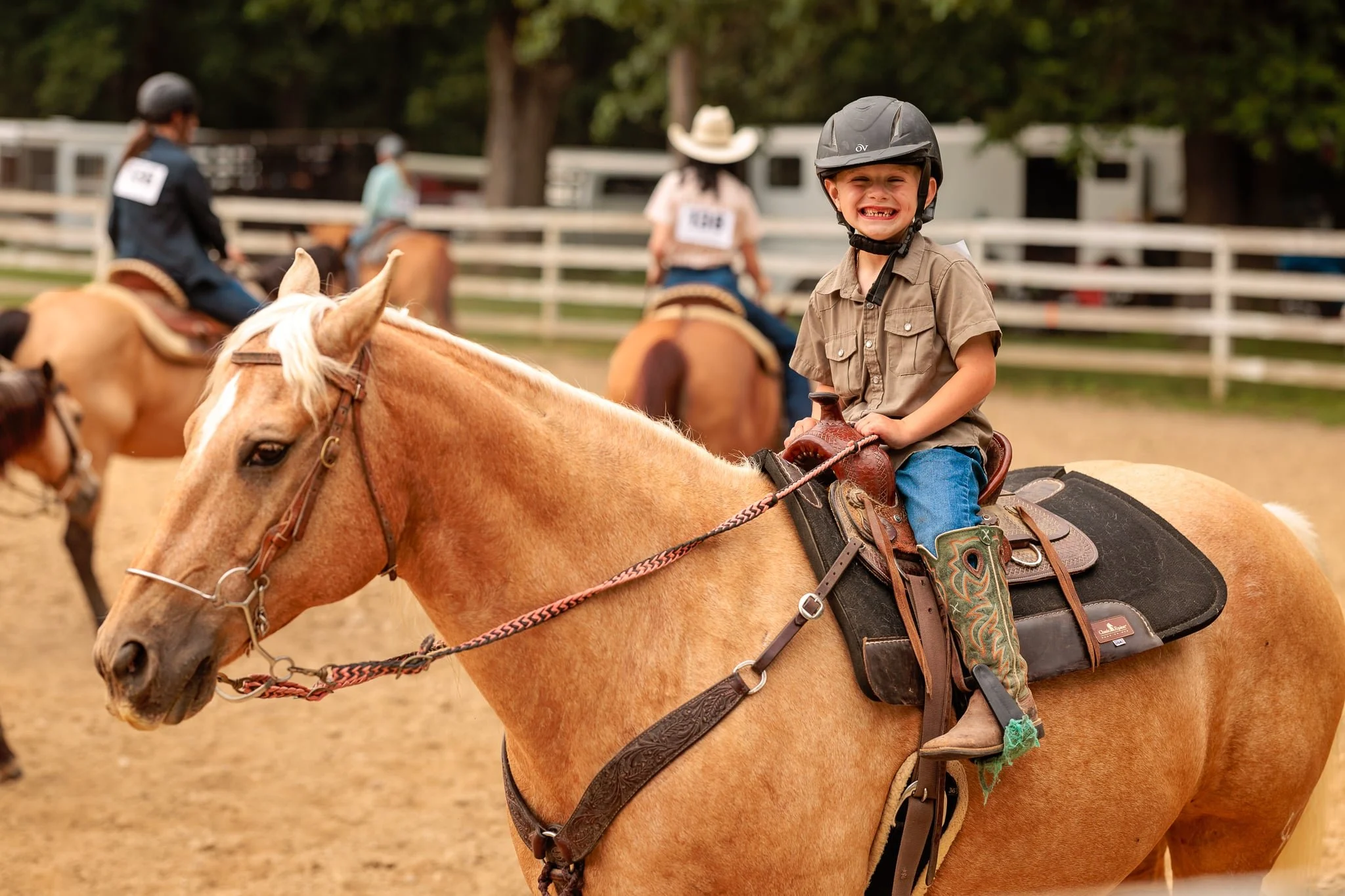 A young boy smiling happily while riding a light brown horse during a horseback riding event, wearing a black helmet and a tan shirt. Other riders and horses are visible in the background, along with a wooden fence and trees.