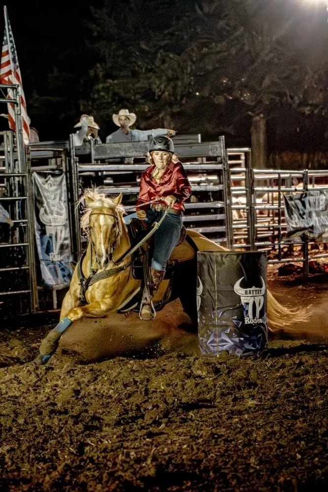 A woman riding a rodeo barrel racing horse at night in an arena, wearing a red shirt and helmet, with a cowboy and another person in the background.
