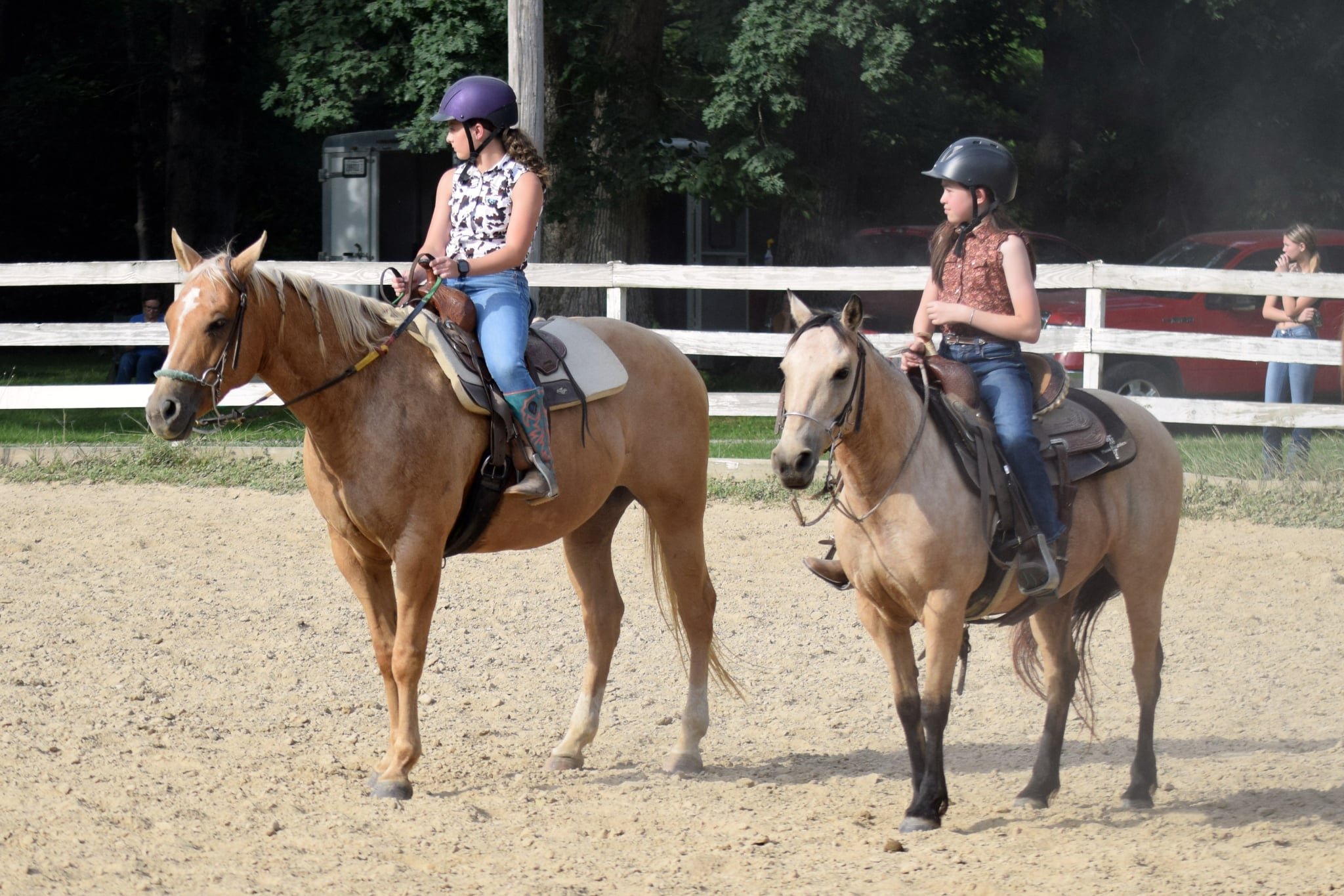 Two young girls riding horses in an outdoor riding arena, wearing helmets and casual clothing, with a wooden fence, trees, and a few onlookers in the background.