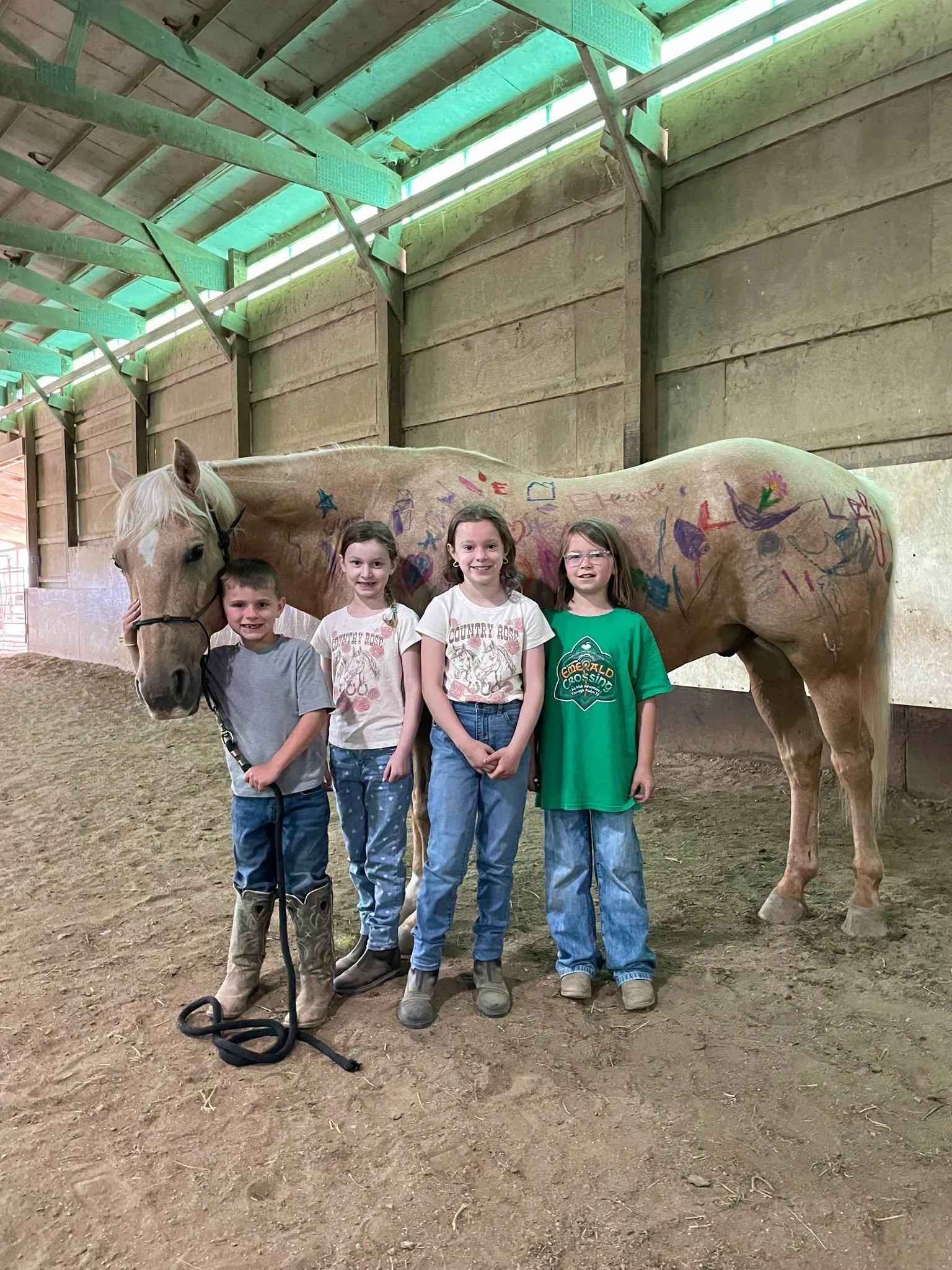 Four children standing in front of a light-colored horse with colorful drawings on its body inside a barn. The children are smiling and wearing casual clothes.