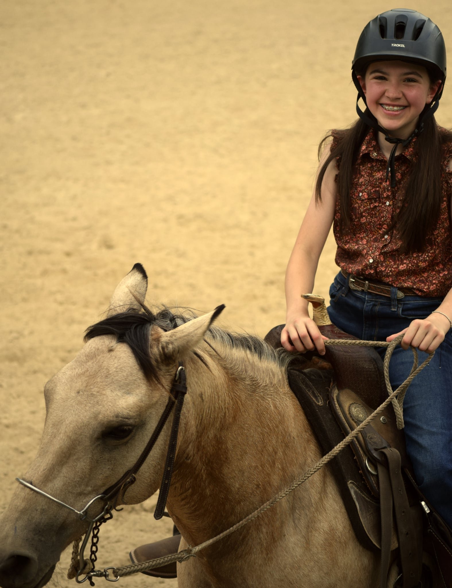 A young girl with braces smiling while riding a horse, wearing a black helmet and a floral sleeveless top.