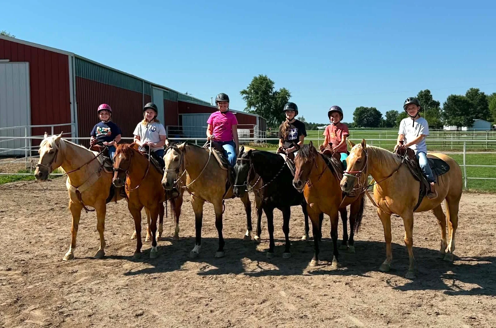 Six children riding horses on a dirt riding arena on a sunny day, with a red barn and green fields in the background.