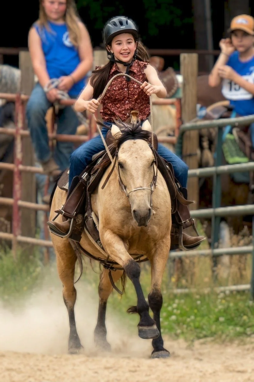 A young girl wearing a helmet rides a galloping light-colored horse during a rodeo event, with spectators watching in the background.