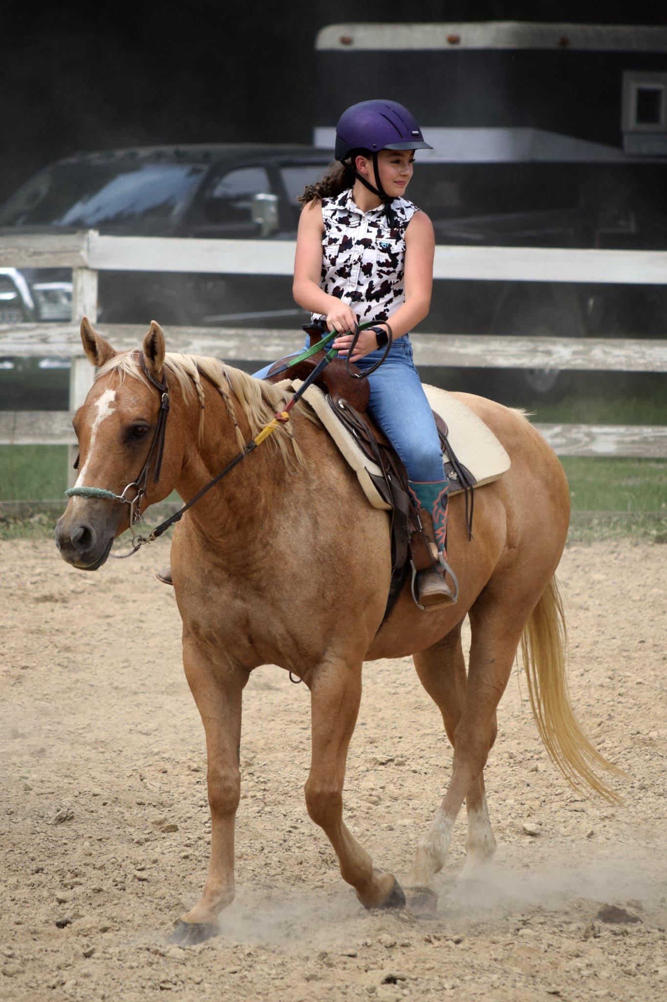 Young girl riding a light brown horse with a blonde mane, wearing a purple riding helmet, cow-patterned sleeveless shirt, and blue jeans in an outdoor riding area with a wooden fence and vehicles in the background.