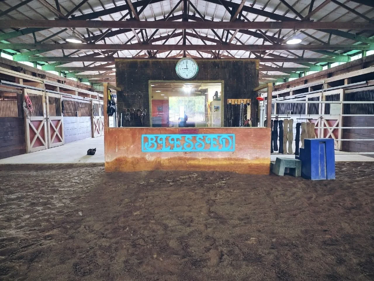 Indoor horse stable with a central reception area displaying a clock and sign, surrounded by horse stalls with grooming tools and equipment.