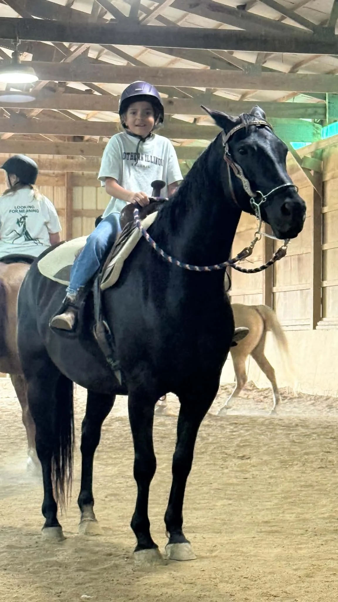 A young boy wearing a helmet riding a black horse in an indoor riding arena. There is another child in the background with a ponytail and a white t-shirt.