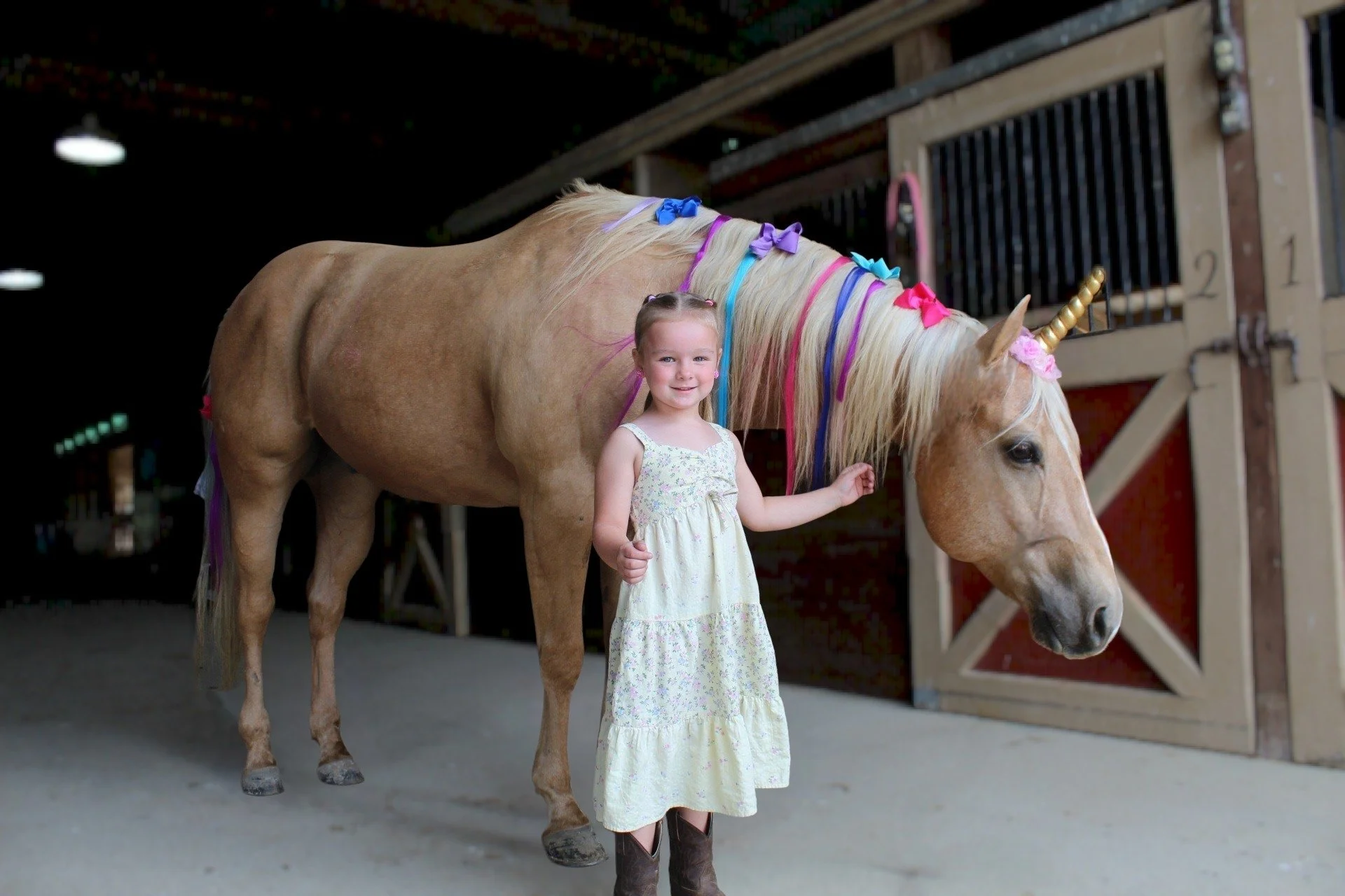 A young girl standing next to a light-colored pony with a rainbow-colored mane decorated with bows. The girl is smiling and touching the pony's mane inside a stable or barn.