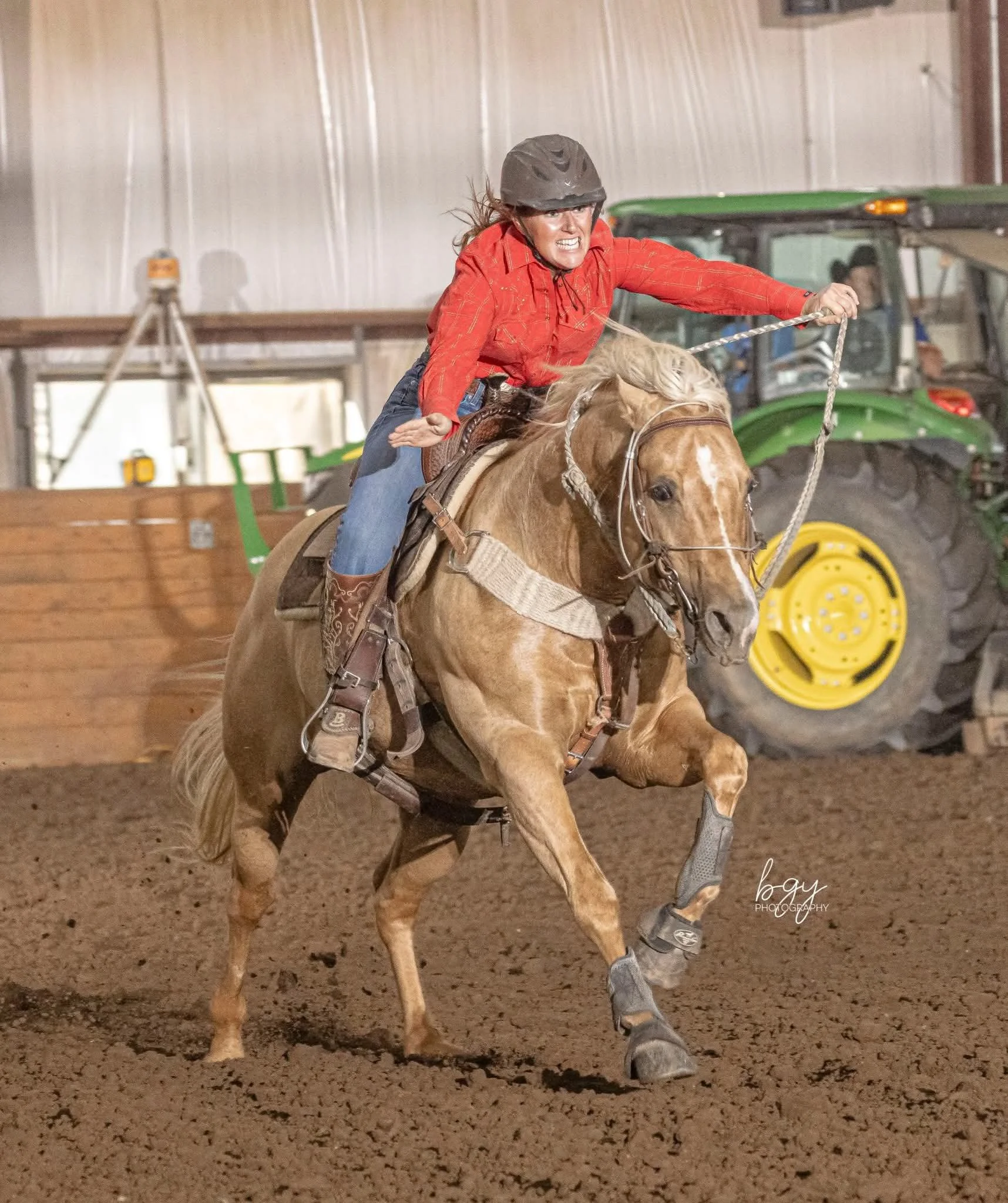 Woman wearing a helmet and a red plaid shirt riding a light brown horse inside a barn, with a green tractor in the background.