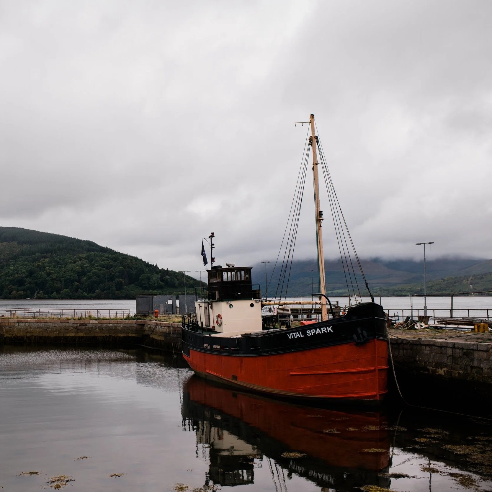 A boat named 'Vital Spark' docked at a harbor with mountains and cloudy sky in the background.