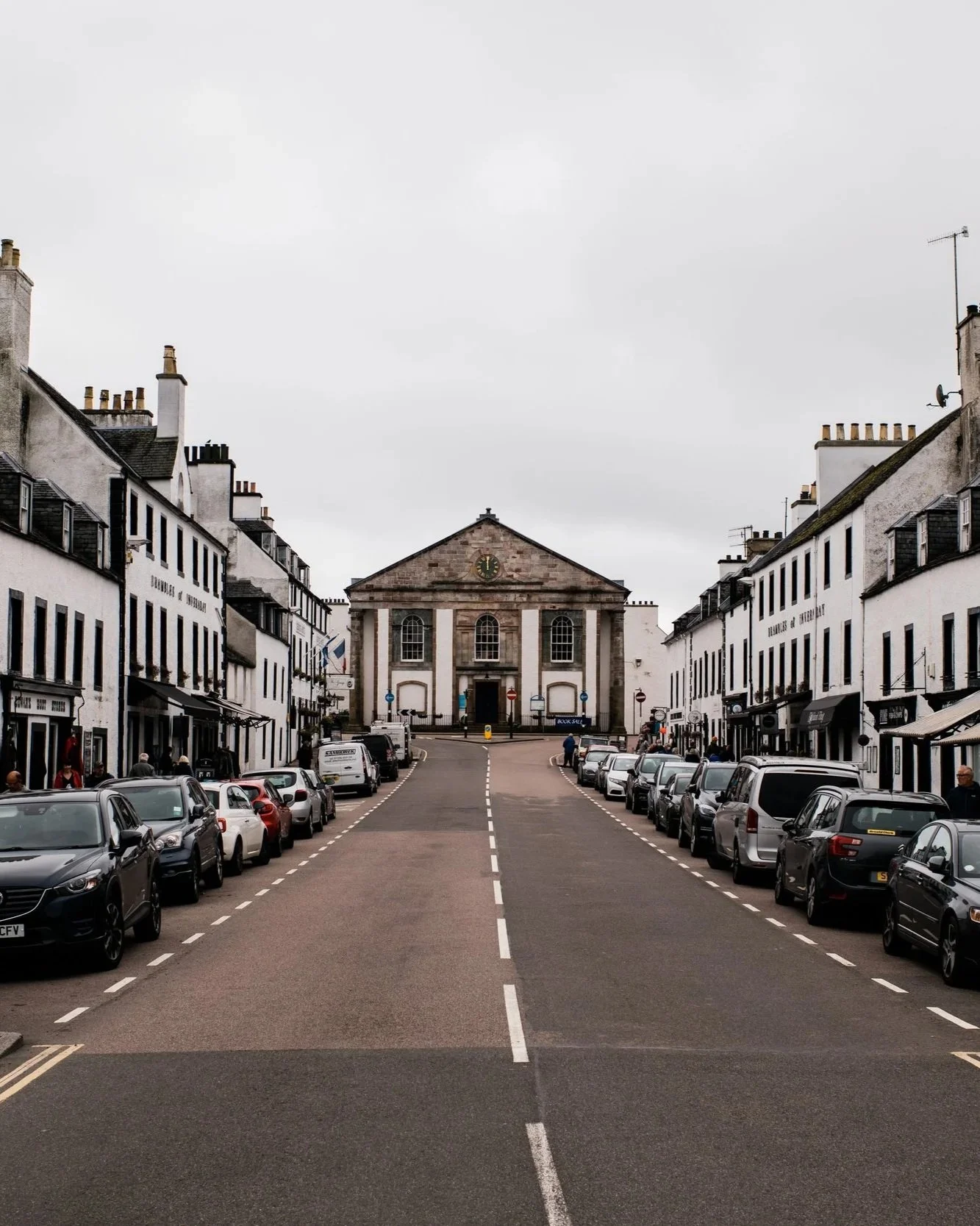 Street view of a small town with parked cars along the road, white buildings with black accents on either side, and a central town hall or church with a clock tower at the end of the street under cloudy skies.