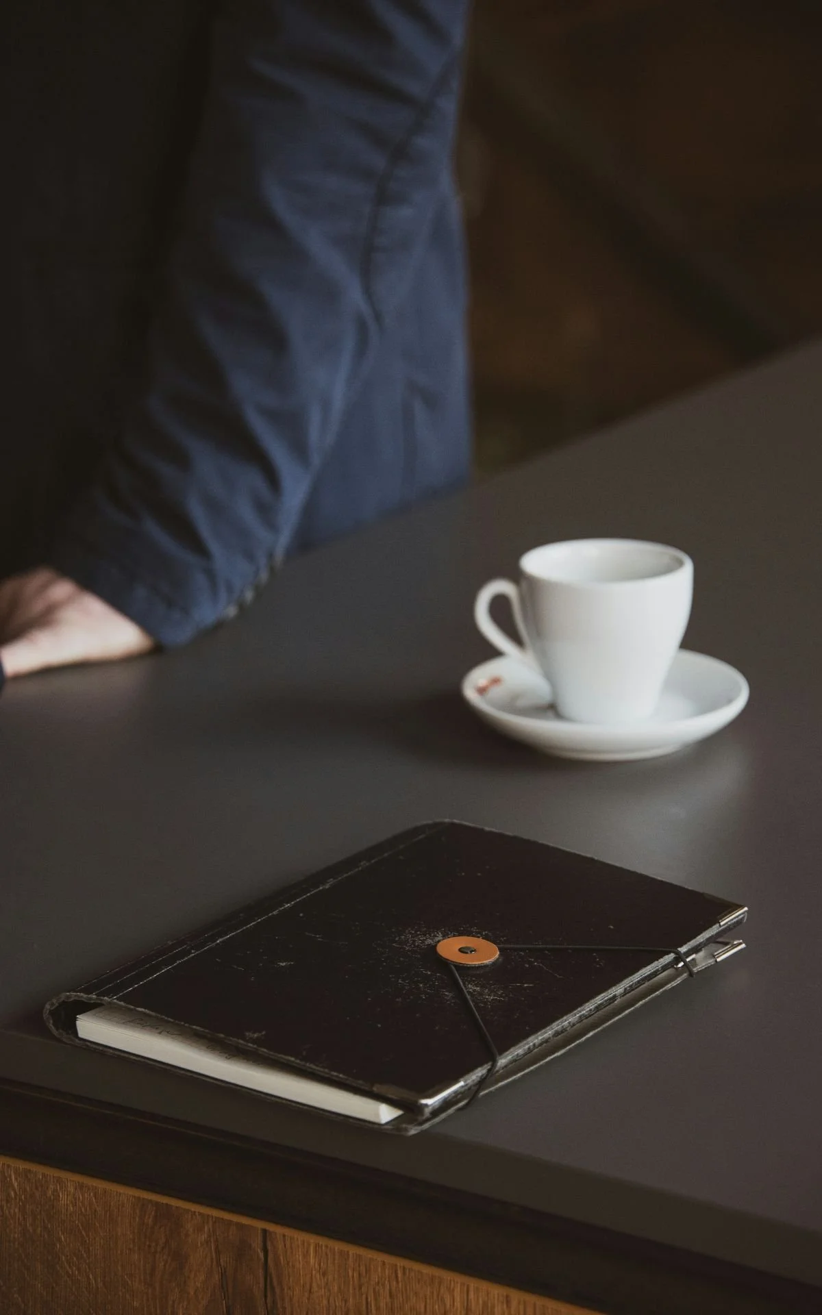A black table with a closed black notebook or journal, a white coffee cup with a matching saucer, and part of a person's arm and hand wearing a blue shirt. The background includes a dark wall and wooden surface.