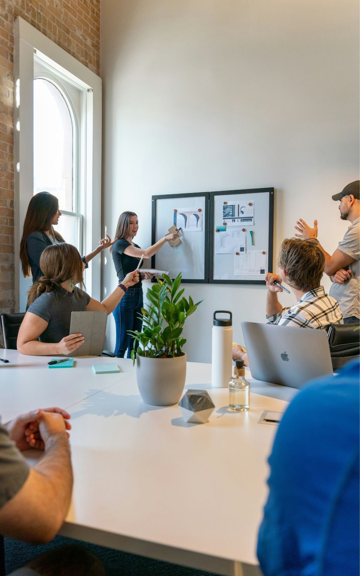 Group of six people in a meeting room with presentation boards on the wall, discussing a project. Some people have laptops or tablets, and a large window lets in natural light.